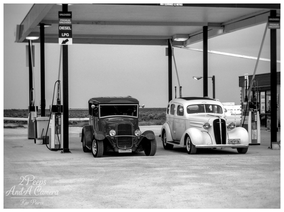 Two vintage cars, possibly hot rods, are parked side by side underneath the awning of the Nullarbor Roadhouse gas station.

The photo is in black and white, giving it a classic, moody feel. The car on the left is dark with a chopped roof, and the car on the right is light-colored and has a slightly older, rounded sedan body.

Fuel pumps (Unleaded, Premium Unleaded, Diesel, LPG) are visible on both sides. The ground is dusty, and the vast, flat landscape of the Nullarbor Plain stretches into the background.