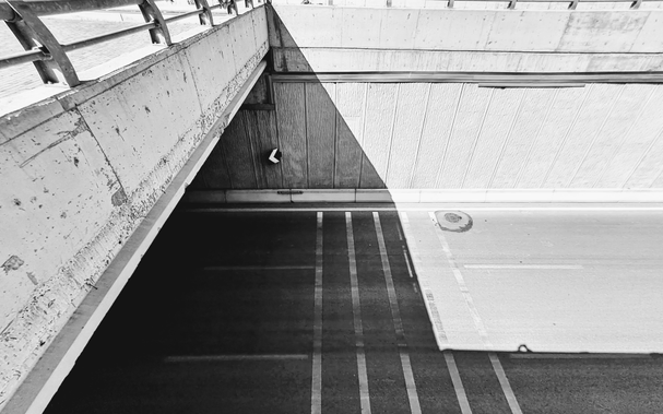 Picture of concrete underpass structure casting sharp triangular shadow diagonally across two-lane roadway with white painted dashed lines in shadow and solid white line in bright area, structure shows exposed concrete walls and metal railing along top left edge; small directional arrow sign visible on dark wall inside shadow on center left of frame