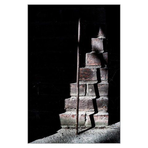 An iron bar is leaning against a wall in a recess in the wall of a house, casting a zigzag shadow on the stepped wall of the chimney. A highly contrasting, dark-toned image.