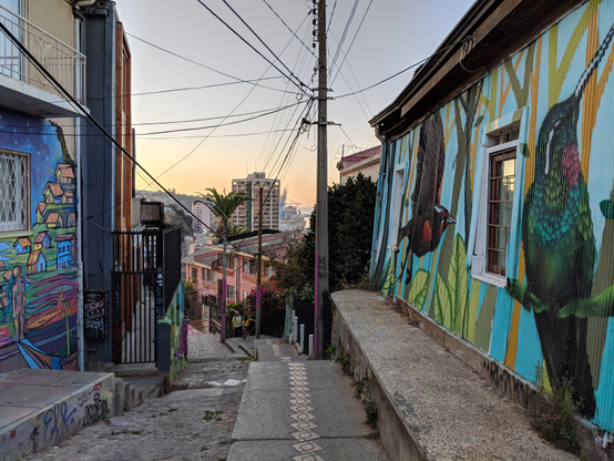 Français : L'image montre une vue plongeante sur un escalier à Valparaíso, au Chili. Les murs de chaque côté sont recouverts de fresques représentant des maisons colorées à flanc de colline et un colibri à gorge rubis. L'escalier serpente entre des clôtures de cour et des bâtiments aux couleurs vives. Au loin, on aperçoit les contours du Cerro Concepción et du port de Valparaíso.

تُظهر الصورة منظرًا لدرج في مدينة فالبارايسو، تشيلي. تغطي الجدران على جانبي الدرج لوحات جدارية تصور منازل ملونة على سفح تل، بالإضافة إلى طائر طنان ذي حلق ياقوتي. يلتف الدرج بين أسوار الأفنية والمباني المطلية بألوان زاهية. في الأفق، تظهر معالم تلة سيرو كونسبسيون وميناء فالبارايسو.
