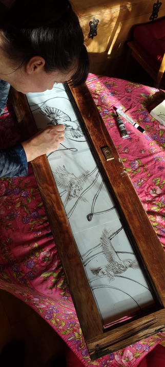 A top-down view of a long, narrow, wood-framed window lying on a table with a pink floral cloth. The window has three thrushes and a series of horizontal curved lines painted on it, outlined in only black. In the top left corner of the image, the top of a woman's head can be seen, with her hair pulled back in a ponytail, and her arm reaching over the window, painting the black lines with a small tube of Pēbēo Cerne Relief (imitation lead).
