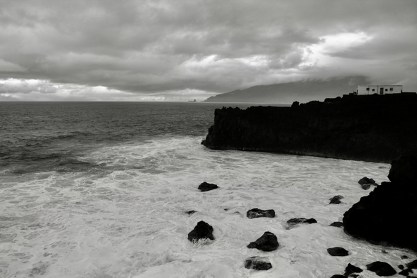 das schwarz-weiß-bild zeigt eine lebhafte meereslandschaft unter einem bedeckten himmel. im vordergrund sieht man eine zerklüftete küstenlinie, an der sich die wellen brechen und weißen schaum erzeugen. einige dunkle felsen ragen aus dem wasser.
im mittelgrund erstreckt sich das offene meer bis zum horizont, wo sich eine unscharfe landmasse unter dem grauen himmel erahnen lässt. am rechten rand der küstenlinie steht ein helles gebäude, vermutlich ein haus.
der himmel ist voller dichter wolken, die eine düstere stimmung erzeugen. das gesamte bild vermittelt ein gefühl von kraft und ungestümheit der natur.