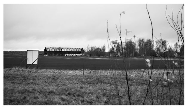 En liggande svartvit bild på en lada vars tak är ett öppet skelett. I förgrunden åker, en bortvänd reklamskylt och lite sly. Till höger om ladan är en dunge träd och ett boningshus. Himmelen är molnig.