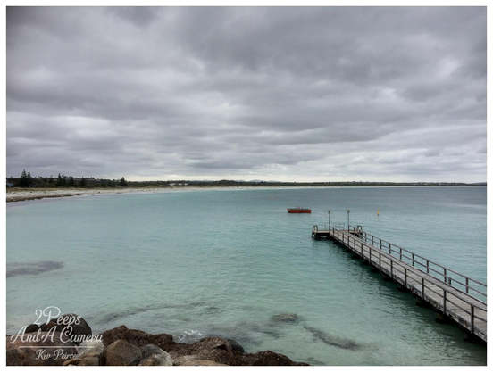 A wide angle landscape photo of a calm turquoise coloured ocean with a long wooden jetty extending from the right foreground.

The jetty leads out toward a small red object floating in the water. The beach runs along the left side of the frame, backed by low coastal scrub and houses under a heavily clouded, grey sky.