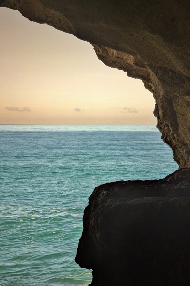 A vertical photograph taken from inside a dark coastal cave, framing a view of the ocean and the sky. The foreground is dominated by the dark, textured rock of the cave mouth, creating a natural frame on the right and top. The mid-ground shows calm, turquoise-blue ocean water extending to the horizon. The sky is a pale, soft gold and beige, suggesting sunrise or sunset. The contrast between the dark cave and the brightly lit sea and sky creates a dramatic and enclosed atmosphere.