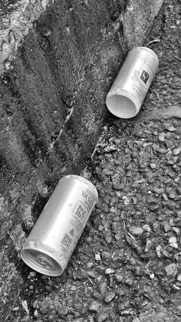 Schwarz-Weiß-Foto von zwei Aluminiumdosen, die im Rinnstein liegen. Sie liegen hintereinander und haben einen Abstand zueinander. 