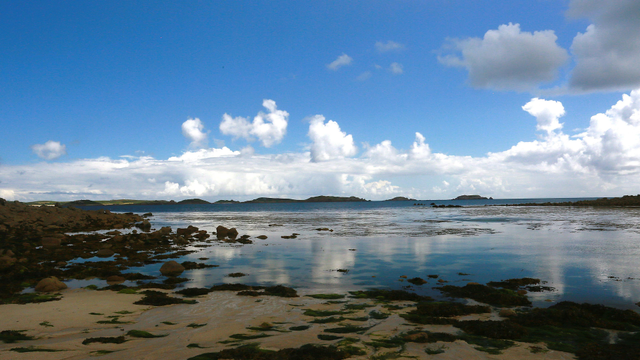 A sandy beach with seaweed in the foreground, lichen-covered rocks to the left, a still sea, reflecting sky and clouds, in the middle ground, and hilly islands in the distance.
