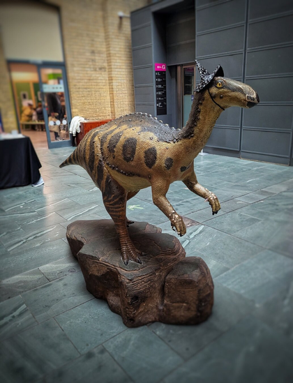 A life-sized model of a dinosaur sculpture displayed indoors on a textured rock-like base. The dinosaur is adorned with a small, black witch’s hat perched on its head, adding a whimsical touch. The sculpture features a detailed, textured body with dark spots and stripes along its back and tail. The setting appears to be an indoor public space, such as a museum or exhibition area, with a tiled floor and glass doors in the background. The scene is softly lit, highlighting the playful contrast between the prehistoric creature and the festive hat.