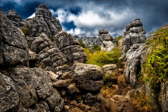 Farbfotografie einer dramatischen Karstlandschaft, aufgenommen auf einem schmalen Pfad zwischen massiven Felsformationen.

Im Vordergrund füllen grosse, horizontal geschichtete und abgerundete Felsbrocken das Bild, links und rechts einen steinigen Pfad freilassend. Die Steine sind grau mit rötlich-braunen Akzenten.

Im Mittelgrund erheben sich imposante, säulenartige Felsformationen. Links dominiert ein hoher, spitz zulaufender Felsgipfel. Rechts und in der Mitte stehen weitere, gestapelte Felsblöcke, die von Büschen und gelb-grüner Vegetation umgeben sind.

Der Hintergrund zeigt eine weite Reihe ähnlicher, zerklüfteter Felsformationen, die sich bis zum Horizont erstrecken.

Der Himmel ist extrem dramatisch und kontrastreich, mit tiefblauem Himmel, der von grossen, weissen und dunklen, schweren Wolken durchzogen wird. Die starke Beleuchtung und Sättigung erzeugen eine intensive, fast surreal wirkende Stimmung.