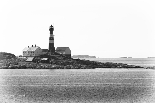 This is a black and white photo in landscape format of the Landegode Lighthouse in Norway's arctic north (2015).

The lower quarter of the image is filled with smooth and calm dark sea water with only small ripples from the afternoons breeze disturbing the surface. To the left of centre is a tall, tapered, cylindrical, lighthouse set on a small and rocky island that stretches from the left and slopes downwards into the sea. Surrounding the lighthouse are a few white, two story houses, a large barn like shed and a few smaller outhouses. our lighthouse is painted a dark colour but with two thick white horizontal stripes about half way up. There are five thin vertical shaped windows facing the camera and the light can be seen shining through. In the distance there a number of small islands, islets and skerries, perhaps a few km away. Above is a hazy, clear and cloudless sky. 

This lighthouse built in 1902 from cast iron and is 29 metres tall. The light is active from dusk to dawn from 4 August until 2 May each year. The light is not active in the summer due to the midnight sun in the region.