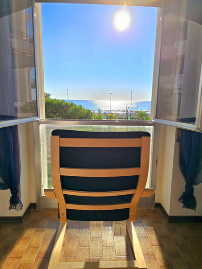 Open window on a warm day, with a comfortable chair positioned to enjoy a breathtaking sea view. Bright sunlight illuminates the azure water and lush coastal vegetation. Curtains on either side of the window sway gently in the sea breeze, creating a relaxing atmosphere and inviting one to immerse in the tranquility of the Mediterranean landscape. Golden sunlight reflects on the tiled floor, emphasizing the warmth and serenity of the setting. The scene evokes a sense of peaceful contemplation and holiday relaxation