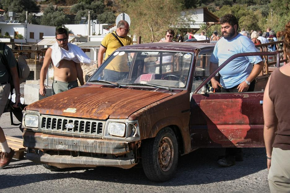 A group of people standing around an old and rusty car with one person entering through the open driver's side door. Houses and trees in the background.