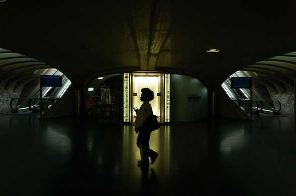 The image captures a silhouetted figure walking through a dimly lit, modern architectural space, a subway station. The dominant color scheme is dark, with deep blacks and shades of gray, creating a dramatic and mysterious atmosphere.

The central focus is the silhouette of a person, walking from left to right. She is carrying a bag over her shoulder and appears to be holding a phone or small device in her hand. Her afro-style hair is clearly defined against the lighter background. 
