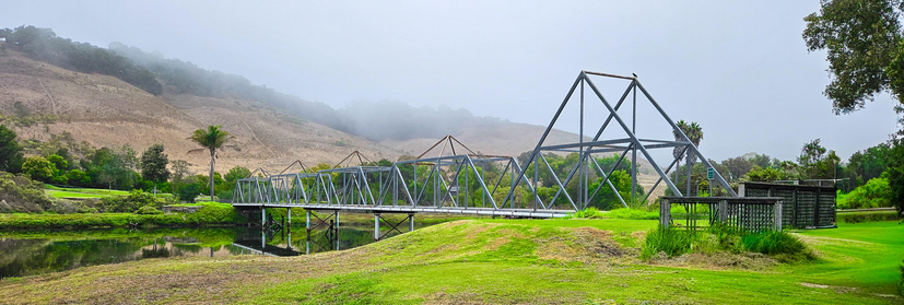 A panoramic view of a metal truss bridge spanning a body of water, set against a backdrop of a green, sloping hillside shrouded in mist. The bridge is constructed of interconnected gray metal beams forming a repeating triangular pattern, and is elevated above the water surface with visible support structures. To the right of the bridge, a small, wooden structure, resembling a simple shed or viewing platform, is partially visible in the foreground. The surrounding landscape features a well-maintained green lawn, with trees lining the edges of the water and the hillside. The overall lighting is soft and diffused, suggesting an overcast day.