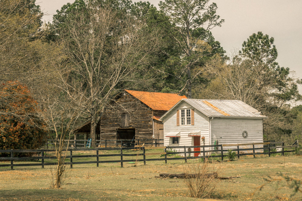 Rural farm scene showing a collection of connected buildings including a weathered dark wooden barn with horizontal siding on the left, and a white painted two-story structure with a tall narrow window on the right. The structures feature rusted orange and silver metal roofs at varying angles. A black wooden post-and-rail fence runs across the foreground of the property, enclosing a sparse grass pasture. Bare deciduous trees and evergreen pines frame the buildings against an overcast sky. The property shows signs of age and weathering, with some deterioration visible on the structures. A small tire swing hangs on the rightmost building, and various farm equipment or materials are visible around the property.