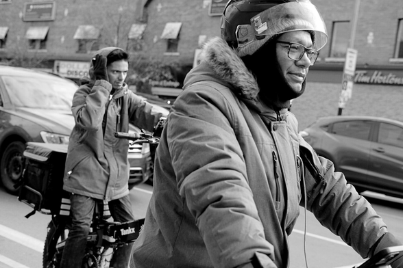The shot is sharply tilted down at the right, on an uneven angle. It is a close shot on two food couriers waiting for the light to change. The one at right, closest to camera and filling the frame, has a parka on and is wearing a helmet with the visor up. The one at left, in the background, has his hand up to his ear.