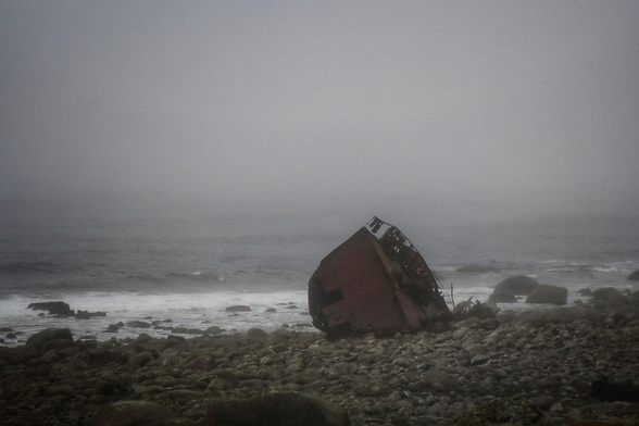 A rusty part of a shipwreck on a stony beach. Its foggy and the colours are almost monochrome