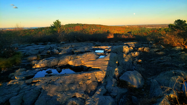 A full moon appears in the late day sky as shadows begin to climb up Chickatawbut Hill which is awash in hues of orange and red.