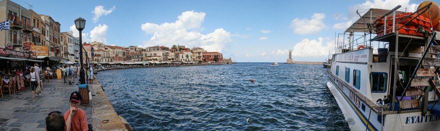 Harbor scene with a boat on the right, a row of buildings with shops and restaurants on the left, a street with people walking, and open water leading to a distant lighthouse.