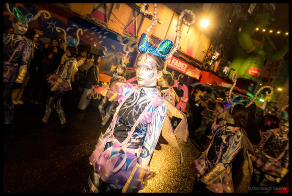 Young dancer from Joan Denise Moriarty School in striking purple, pink and silver painted costume with blue metallic headdress and golden textured arm pieces poses during Dragon of Shandon parade on North Main Street, Cork, with crowd and colourful murals in background.