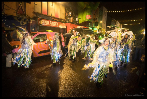 A group of dancers in flowing white and blue costumes with green netting, feathers and LED lights performing on wet North Main Street, Cork, during the Dragon of Shandon parade at night with King Fadez shop and string lights visible.
