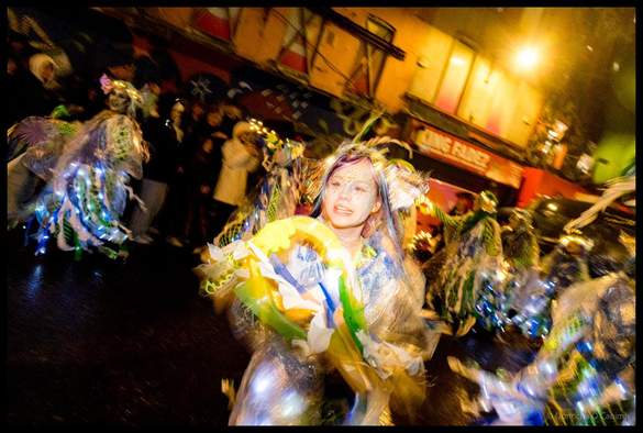 Motion-blurred photograph of a dancer in elaborate yellow, white and green costume with flowing fabric and LED lights spinning during Dragon of Shandon parade performance on North Main Street, Cork, at night.