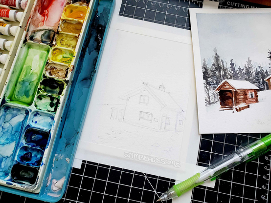 A photo of an in progress artwork of a cabin, currently drawn in graphite with loose lines and details that will be painted in and refined later. Next to the artwork is another piece, this one a completed watercolor painting of a cabin in winter with snow and trees around it. There is also watercolor paints and a pencil in frame.