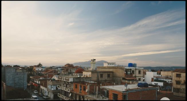 A quiet townscape under a wide, pastel sky. Old, low-rise buildings with flat rooftops and water tanks spread across the foreground, while a mosque minaret and distant hills are visible in the background. Soft evening or early morning light gives the scene a calm, warm tone.