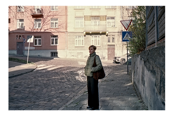 A color film photo capturing a person standing on a sidewalk at a street corner. The street is made of cobblestones. The person has short hair and wears a green quilted jacket, wide-leg dark trousers, and carries a large brown bag. In the background are two apartment buildings, one pink and one beige, under bright sunlight.