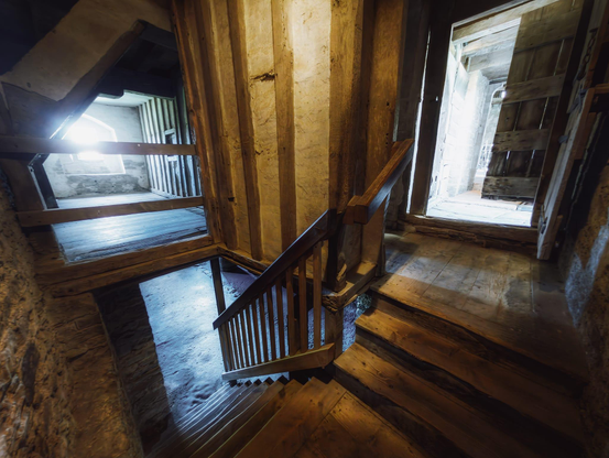 The interior of Stokesay Castle's Solar Block stairwell descends into shadow and history, with substantial timber posts and beams rendered in warm honey and amber tones creating a framework of medieval craftsmanship. Bright daylight streams through two tall windows, one on either side of the staircase, casting sharp geometric shadows across the worn wooden steps and illuminating the rendered stone walls with an ethereal blue-white glow that contrasts strikingly with the aged timber. A sturdy wooden handrail with vertical balusters guides the descent, whilst the exposed ceiling beams and structural supports overhead speak to the 14th-century construction, all contributing to the palpable sense of stepping back through centuries as one moves from the comparatively brighter ground floor towards the dim cellar below.
