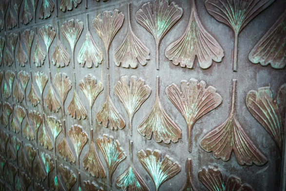 A close-up photograph shows a textured, metallic surface covered in repeating ginkgo leaf-shaped patterns. The surface is dark, likely bronze or a similar alloy, with patches of green oxidation visible throughout. Each leaf is three-dimensional, raised from the background and appears to have a central stem or vein. The leaves are arranged in neat rows and columns, forming a grid-like pattern across the entire frame. The lighting casts subtle shadows, enhancing the depth and detail of the embossed design.

Provided by @altbot, generated privately and locally using Gemma3:27b