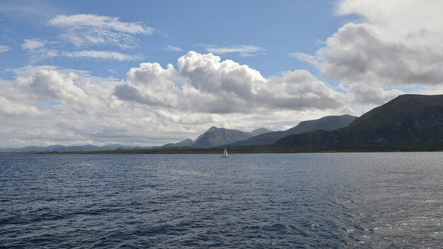 A photo of water leading into mountains. There is a sailboat in the water. The sky is blue with white clouds in it.