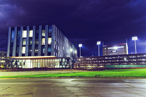To the left side of the frame a rectangular building has some of the windows out of four stories lit,  to the center and the right a concrete parking garage and the lights of a sports stadium