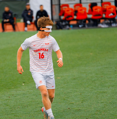Number 16 wears a white mask and a white shirt with red lines on a mostly empty green field with  people on the sidelines behind.