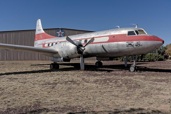A color landscape photo of an old, twin-engine propeller driven airplane. It is silver with a red stripe. It is a commercial airliner and it sits on tricycle landing gear. A hanger is behind the plane. It is a static display on a dark-red lava rock / dry grass field. The sky is blue. 