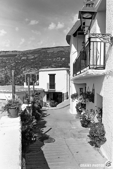Narrow concrete street in Capileira, a Mediterranean village lined with white buildings, balconies, and flower pots. Hills and mountains rise in the background beneath a partly cloudy sky.