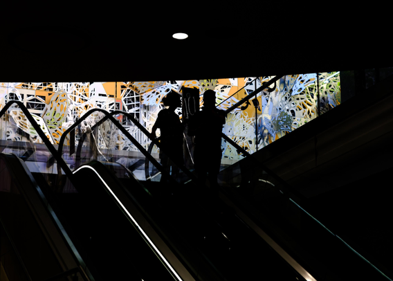 High contrast low key picture of a couple going down an escalator.  In the background is a colourful brightly lit on a sunny day