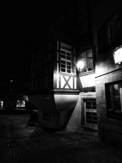 Schwarzweißfotografie einer nächtlichen Straßenszene vor einem Fachwerkhaus in einer Kleinstadt. Das Haus ist rechts im Bild. Durch eine Laterne werden links ein hervorspringender Erker und rechts ein Teil der Fassade mit diversen Sprossenfenstern beleuchtet. Alles andere verschwindet mehr oder weniger in der Dunkelheit der Nacht. 