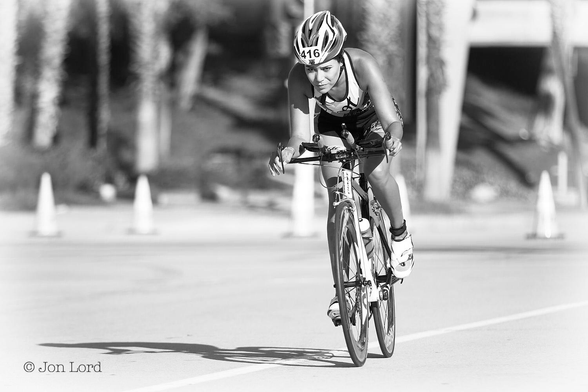 This is a black and white sports photo in landscape format of a young woman in a cycle road race. Long Beach, California (2015).

Very slightly to the right of centre is a young woman riding a sports bike towards the camera and from right to left. Our rider has a look of determination and concentration on her face. She is isolated with no other competitors visible. The rider is bent forward over the handle bars, is looking ahead with her head bend downwards. She is wearing a cycle helmet with the number 416 on the front in bold black numerals, a sleeveless t-shirt and shorts. The bike is in a slight bank to the right having completed a steep bend in the road. The woman's and her bike's shadow is clear and about the same size on the rider's right side. The background is blurred out of focus but road cones marking the track are visible in the near distance as is a tree lines embankment.