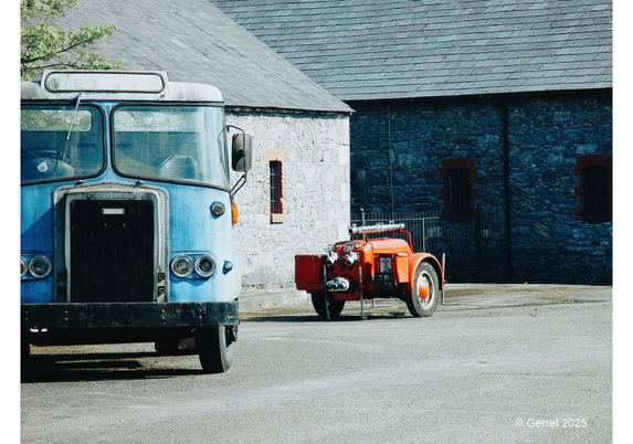 A vintage blue truck and a red fire engine are parked on a paved area in front of a stone building. The scene conveys a nostalgic, historical atmosphere.