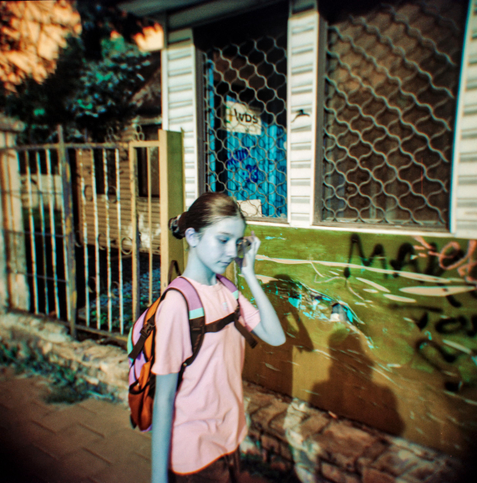 A young girl with a backpack walks along a sunlit street, her hand raised to her face in a thoughtful gesture. The scene is rendered in surreal turquoise and amber tones, giving her skin a cool blue tint against the warm hues of the wall and metal fence behind her. The dreamy color shift and slight blur characteristic of the Holga create a nostalgic, almost cinematic atmosphere — as if capturing a fleeting memory from another world.