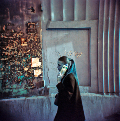 A woman walks past a textured urban wall, holding a smartphone to her ear. The tones are inverted and ethereal — her hair and face appear bluish, while the background glows in muted teal and magenta shades. The rough surface of the wall and scattered posters contrast with the modern device she’s holding, emphasizing a tension between past and present. The Holga’s soft focus and LomoChrome Turquoise film transform the ordinary moment into something surreal and introspective.