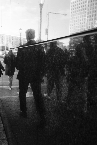 A group of people are seen through a reflection on a marble surface. They are all walking on the sidewalk in the same direction, away from the camera. In the background a couple of buildings and street lights puncture the sky. The person in the foreground is masculine and wearing a suit.