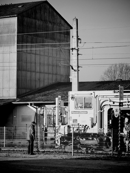 Das Bild ist in Schwarzweiß gehalten und zeigt eine Szenerie im Bahnhofsbereivh. Im Vordergrund steht ein Mann mit Mütze und dunkler Jacke neben einem Zaun, der zu einer Maschine blickt. Die Maschine trägt die Aufschrift “Plasser &amp; Theurer” und ist ein gleisgebundenes Wartungs- oder Baugerät für den Schienenverkehr. Dahinter sind ein Industriegebäude mit Wellblechfassade, ein Strommast mit zahlreichen Oberleitungen sowie Teile eines niedrigeren Gebäudes zu sehen. Im Hintergrund ragen die kahlen Äste eines Baumes ins Bild. Die gesamte Komposition vermittelt eine technische und zugleich ruhige Atmosphäre, betont durch den Kontrast von Licht und Schatten.