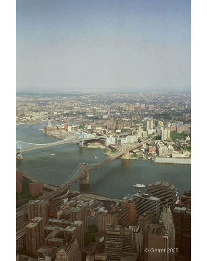 Aerial view of a cityscape featuring two large suspension bridges spanning a river, surrounded by urban buildings under a hazy, blue sky.