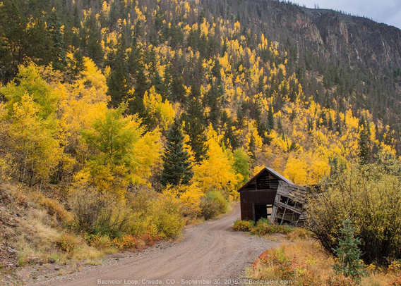 A dirt road curves around an old wood building through a valley with a mix of green conifer and golden aspen trees; some cliffs in the distance; tiny slice of grey blue sky in upper right.
©BosqueBill.com
