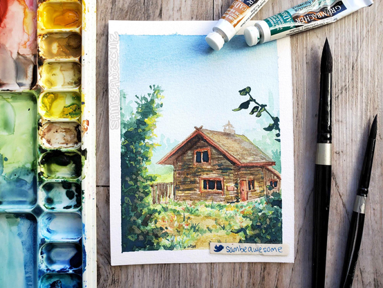A photo of a watercolor painting of a wooden cabin surrounded by greenery and nature with a bright clear blue sky. Paints and brushes lay next to the artwork.