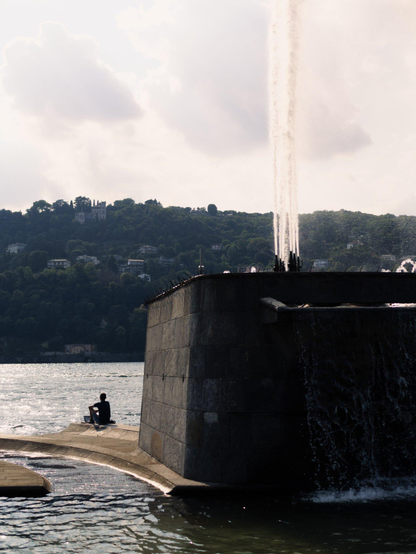 A young man sits on a smooth concrete structure near the lakefront. On his side is a large concrete fountain with a tall water jet. We only see his silhouette as he's back-lit by bright sunlight.