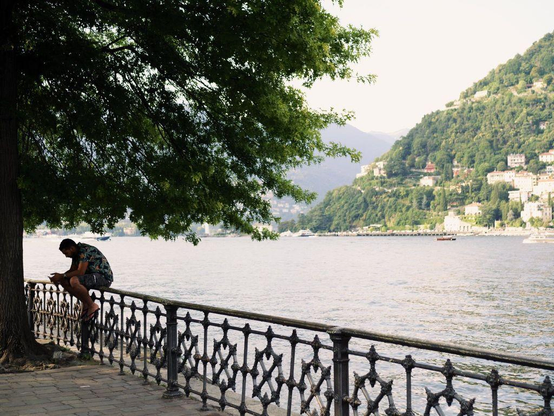 A man sits on a wrought iron balustrade under a tree on the lakeside. In the distance is the opposite side of the lake with mountains and buildings.