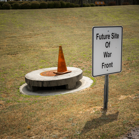 A darkly humorous outdoor installation on patchy grass featuring a white rectangular sign with black text reading "Future Site Of War Front" mounted on a metal post. Next to it stands a circular concrete structure resembling a utility access point or manhole cover, topped with a weathered orange traffic cone. The scene is set in an open field with hedges and trees visible in the background, creating an absurdist commentary on mundane infrastructure and conflict.